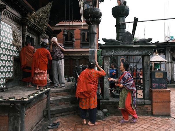 Several individuals stand near an ornate temple, with stone carvings and brick structures visible. The people are dressed in colorful traditional clothing, some appearing to be in prayer or performing a ritual. Two people face each other, seemingly engaged in a conversation or exchange. The setting suggests a cultural or religious significance.