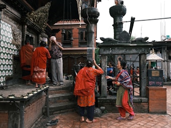 Several individuals stand near an ornate temple, with stone carvings and brick structures visible. The people are dressed in colorful traditional clothing, some appearing to be in prayer or performing a ritual. Two people face each other, seemingly engaged in a conversation or exchange. The setting suggests a cultural or religious significance.