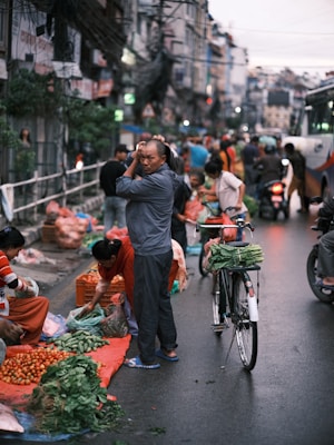A busy street market scene with vendors selling fresh produce. Crates of tomatoes, leafy greens, and other vegetables are displayed on the ground. People are engaged in buying and selling activities, with a bicycle parked nearby carrying leafy vegetables on its rack. Buildings and utility wires are visible in the background, indicating an urban setting.