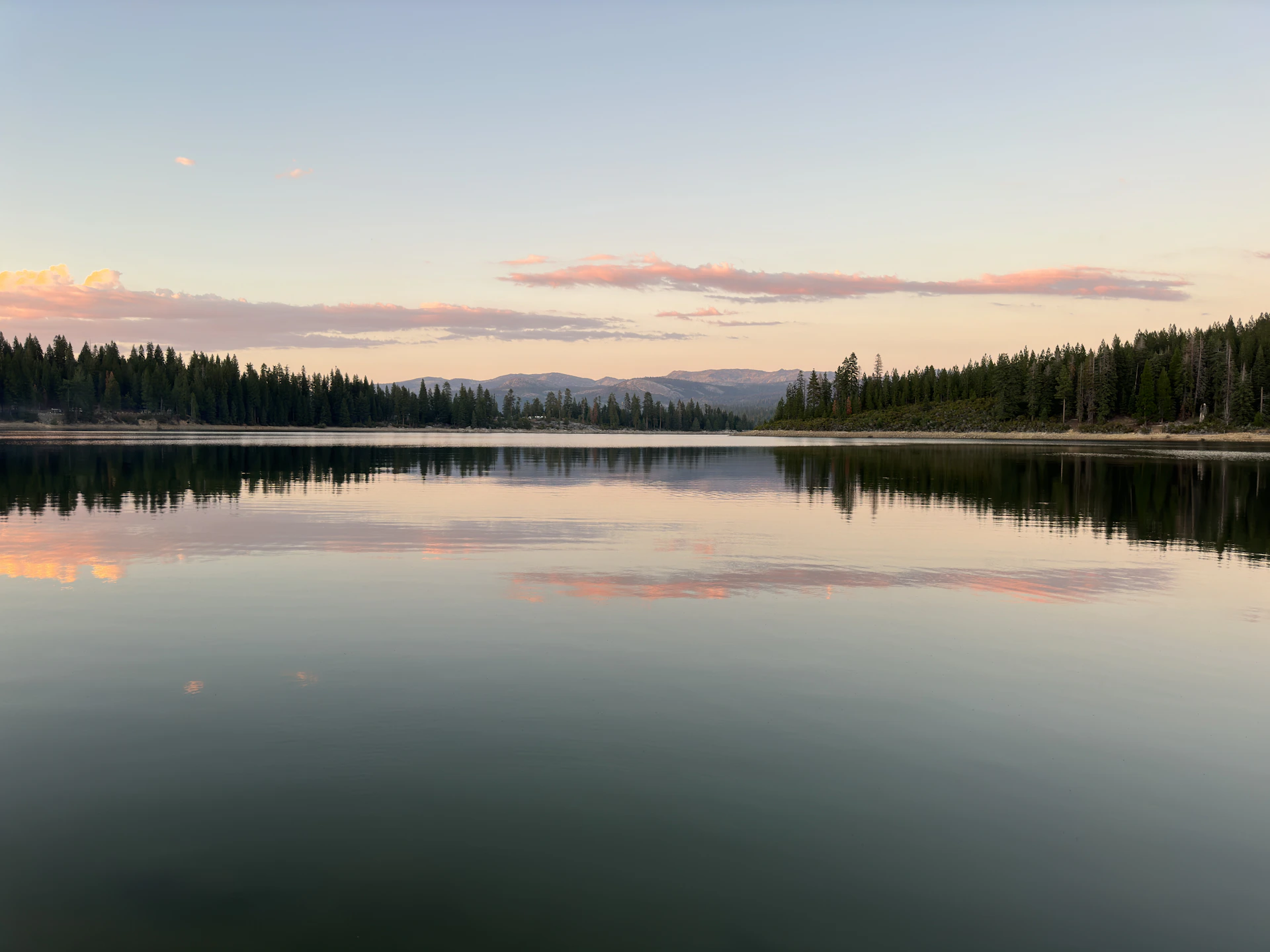 A serene mountain lake at dawn, its glassy surface reflecting the soft pastel sky and surrounding pine trees.