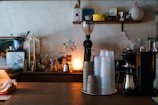 A cozy coffee shop counter featuring a variety of coffee equipment, including a coffee grinder, stacks of disposable cups, coffee brewers, and a small lamp casting warm light. Shelves in the background hold various coffee-related items and decorations.