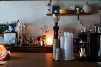 A cozy coffee shop counter featuring a variety of coffee equipment, including a coffee grinder, stacks of disposable cups, coffee brewers, and a small lamp casting warm light. Shelves in the background hold various coffee-related items and decorations.