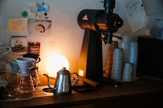 A cozy coffee bar setup featuring a coffee grinder, a metallic kettle, and a drip coffee maker positioned on a counter. The background is adorned with small posters and stickers, and a warm light emits a soft glow, enhancing the ambiance. Several stacks of disposable cups are arranged neatly beside the grinder.