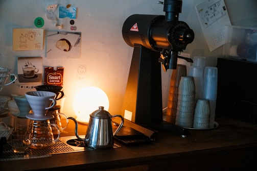 A cozy coffee bar setup featuring a coffee grinder, a metallic kettle, and a drip coffee maker positioned on a counter. The background is adorned with small posters and stickers, and a warm light emits a soft glow, enhancing the ambiance. Several stacks of disposable cups are arranged neatly beside the grinder.