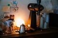A cozy coffee bar setup featuring a coffee grinder, a metallic kettle, and a drip coffee maker positioned on a counter. The background is adorned with small posters and stickers, and a warm light emits a soft glow, enhancing the ambiance. Several stacks of disposable cups are arranged neatly beside the grinder.