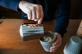 Barista pouring latte art into a ceramic cup on a rustic wooden counter.