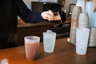 A person is pouring espresso from a small glass pitcher into a transparent cup filled with ice. There are stacks of empty plastic cups in the background, and another cup with a dark beverage already poured sits on the counter.