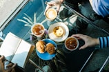 A table with a clear glass top showcases an arrangement of three beverages, including a latte with latte art and two iced drinks. There are pastries on white and blue plates, consisting of a tart and a couple of sandwiches. Two people's hands reach for the drinks, while another person holds an open book. Sunlight casts shadows across the table.