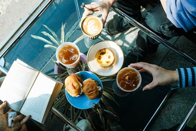 A table with a clear glass top showcases an arrangement of three beverages, including a latte with latte art and two iced drinks. There are pastries on white and blue plates, consisting of a tart and a couple of sandwiches. Two people's hands reach for the drinks, while another person holds an open book. Sunlight casts shadows across the table.