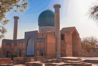 A historic Islamic architectural structure with a large blue dome and intricate geometric patterns on the facade. It features two tall minarets and is surrounded by trees and ancient brick ruins. The overall setting is serene and bathed in warm sunlight.
