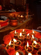 The image depicts a traditional setup with oil lamps (diyas) arranged on a decorative plate, each with a burning flame. Around the lamps, there are marigold flowers and petals. In the background, there are various metallic containers, incense sticks, and religious artifacts placed on an ornately designed table.