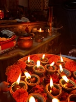 The image depicts a traditional setup with oil lamps (diyas) arranged on a decorative plate, each with a burning flame. Around the lamps, there are marigold flowers and petals. In the background, there are various metallic containers, incense sticks, and religious artifacts placed on an ornately designed table.