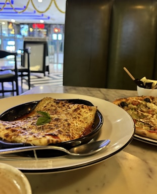 A table setting in a restaurant with a plate of lasagna garnished with a basil leaf. Next to it is a pizza topped with vegetables and olives. There is a cup, possibly containing a hot beverage, partly visible in the foreground, and a bowl with utensils. The restaurant interior has a modern design with a polished marble table and dark green cushioned seating.
