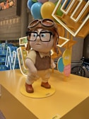 A statue of a cartoon character with oversized glasses, a pilot hat, and a vintage outfit stands on a yellow platform. Surrounding the character are multi-colored balloons and a neon sign. The setting appears to be indoors, with a modern architectural backdrop and a bicycle partially visible in the background.
