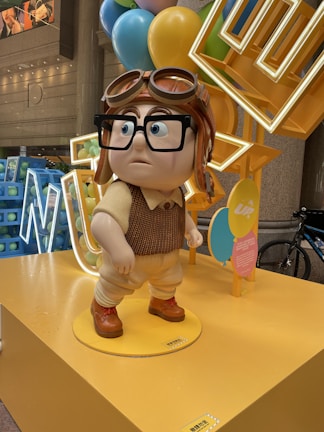 A statue of a cartoon character with oversized glasses, a pilot hat, and a vintage outfit stands on a yellow platform. Surrounding the character are multi-colored balloons and a neon sign. The setting appears to be indoors, with a modern architectural backdrop and a bicycle partially visible in the background.