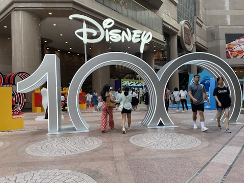 A large silver sculpture forming the number 100 stands in front of a Disney sign at an event space. People are walking around and interacting with the installation. The background includes signage and other decorative elements in a busy urban environment.