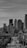 A black and white photograph depicting a cityscape with a focus on skyscrapers. The tall buildings dominate the background, while the foreground features a street with cars and utility poles. The photo captures an urban environment with a slightly overcast sky overhead.