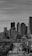 A black and white photograph depicting a cityscape with a focus on skyscrapers. The tall buildings dominate the background, while the foreground features a street with cars and utility poles. The photo captures an urban environment with a slightly overcast sky overhead.