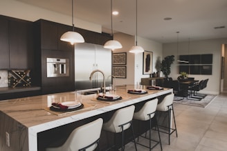 A modern kitchen and dining area with a large marble-topped island featuring elegant bar stools. Pendant lights hang above the island, which is set with plates and burgundy napkins. The kitchen has stainless steel appliances, dark cabinetry, and a wine rack. The dining area includes a rectangular table surrounded by black chairs, with a potted plant and wall art providing decor.