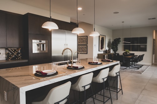 A modern kitchen and dining area with a large marble-topped island featuring elegant bar stools. Pendant lights hang above the island, which is set with plates and burgundy napkins. The kitchen has stainless steel appliances, dark cabinetry, and a wine rack. The dining area includes a rectangular table surrounded by black chairs, with a potted plant and wall art providing decor.