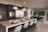 A modern kitchen and dining area with a large marble-topped island featuring elegant bar stools. Pendant lights hang above the island, which is set with plates and burgundy napkins. The kitchen has stainless steel appliances, dark cabinetry, and a wine rack. The dining area includes a rectangular table surrounded by black chairs, with a potted plant and wall art providing decor.
