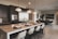 A modern kitchen and dining area with a large marble-topped island featuring elegant bar stools. Pendant lights hang above the island, which is set with plates and burgundy napkins. The kitchen has stainless steel appliances, dark cabinetry, and a wine rack. The dining area includes a rectangular table surrounded by black chairs, with a potted plant and wall art providing decor.