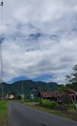 A rural road flanked by lush greenery and small houses. A mountain range is visible in the background under a partly cloudy sky. There are signs and a utility pole along the roadside.