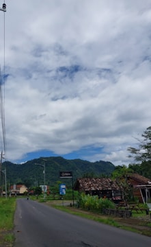 A rural road flanked by lush greenery and small houses. A mountain range is visible in the background under a partly cloudy sky. There are signs and a utility pole along the roadside.