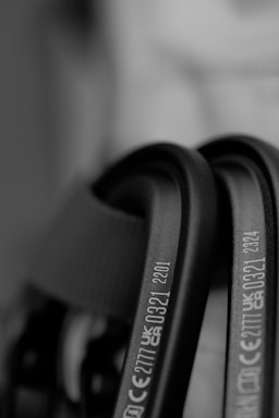 Close-up of black metallic carabiners with engraved CE and UKCA markings. The focus is on the text, providing a clear view of certification numbers and letters, set against a blurred background.