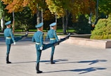 Three uniformed guards are performing a synchronized marching drill in an outdoor setting. The scene is set in a park-like area with green and autumnal trees in the background. The guards wear teal uniforms with matching hats and are holding rifles. Their postures are synchronized, with one leg raised high in unison.