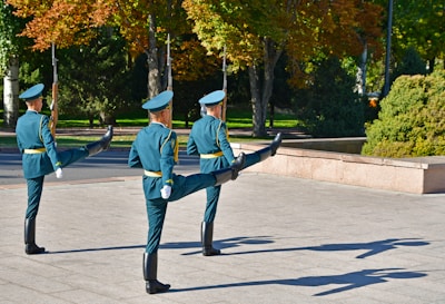 Three uniformed guards are performing a synchronized marching drill in an outdoor setting. The scene is set in a park-like area with green and autumnal trees in the background. The guards wear teal uniforms with matching hats and are holding rifles. Their postures are synchronized, with one leg raised high in unison.