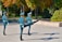 Three uniformed guards are performing a synchronized marching drill in an outdoor setting. The scene is set in a park-like area with green and autumnal trees in the background. The guards wear teal uniforms with matching hats and are holding rifles. Their postures are synchronized, with one leg raised high in unison.