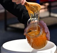 A person is pouring thick, golden honey from a wooden spoon into a jar, captured in close-up. The honey is glossy and richly colored, catching the light as it flows. The person is wearing a dark long-sleeved garment, and the jar is being held over a white container.