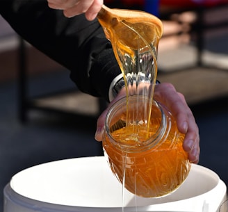 A person is pouring thick, golden honey from a wooden spoon into a jar, captured in close-up. The honey is glossy and richly colored, catching the light as it flows. The person is wearing a dark long-sleeved garment, and the jar is being held over a white container.
