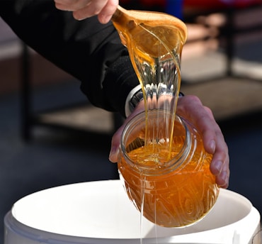 A person is pouring thick, golden honey from a wooden spoon into a jar, captured in close-up. The honey is glossy and richly colored, catching the light as it flows. The person is wearing a dark long-sleeved garment, and the jar is being held over a white container.