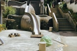A playground with an elephant-shaped slide, complete with eyes and an extended trunk that forms the slide. Surrounding the slide are concrete surfaces and steps leading up an incline, with greenery partially visible. A few children seem to be climbing the stairs, adding a sense of scale and activity.