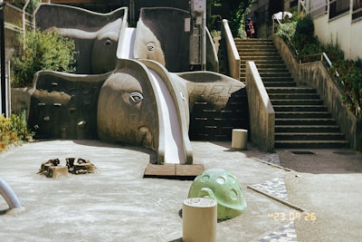 A playground with an elephant-shaped slide, complete with eyes and an extended trunk that forms the slide. Surrounding the slide are concrete surfaces and steps leading up an incline, with greenery partially visible. A few children seem to be climbing the stairs, adding a sense of scale and activity.