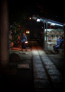 A dimly lit street market scene features a vendor stall adorned with colorful wreaths under fluorescent lights. Beside the stall, a man sits on a chair, while another person with a backpack walks past. The cobblestone path leads to the darker areas of the market, where glowing lanterns create a warm ambiance.