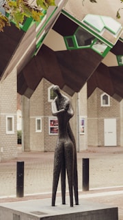 A modern and abstract sculpture stands in an outdoor urban setting. The sculpture is crafted from a dark material, possibly bronze, and depicts a stylized human figure with an elongated form. In the background, there are unique cubical houses featuring geometric windows and differing shades of beige, brown, and green. The contrast between the art piece and the architectural elements creates an intriguing visual composition.