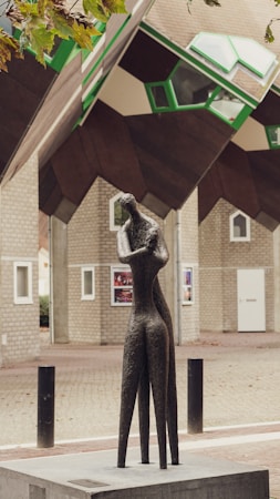 A modern and abstract sculpture stands in an outdoor urban setting. The sculpture is crafted from a dark material, possibly bronze, and depicts a stylized human figure with an elongated form. In the background, there are unique cubical houses featuring geometric windows and differing shades of beige, brown, and green. The contrast between the art piece and the architectural elements creates an intriguing visual composition.