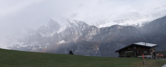 A tranquil mountain landscape features snow-capped peaks partially obscured by mist, creating a dramatic and serene atmosphere. In the foreground, a modern house with a sloped roof rests on a grassy hill, with faint sunlight casting gentle shadows. The scene is enveloped in a quiet, natural beauty, highlighting the contrast between the rugged mountains and the peaceful meadow.