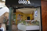 A modern ice cream shop with a storefront that features a curved glass counter displaying various ice cream cones. The shop has an inviting interior with yellow and brown tones, and a prominent wall sign that reads 'The Taste of Happiness'. There are decorative plants outside near the entrance.