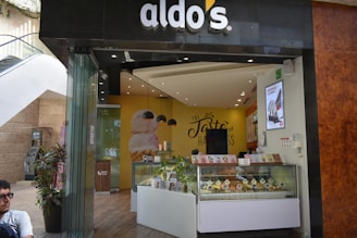 A modern ice cream shop with a storefront that features a curved glass counter displaying various ice cream cones. The shop has an inviting interior with yellow and brown tones, and a prominent wall sign that reads 'The Taste of Happiness'. There are decorative plants outside near the entrance.