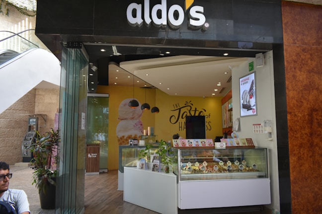A modern ice cream shop with a storefront that features a curved glass counter displaying various ice cream cones. The shop has an inviting interior with yellow and brown tones, and a prominent wall sign that reads 'The Taste of Happiness'. There are decorative plants outside near the entrance.