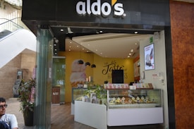 A modern ice cream shop with a storefront that features a curved glass counter displaying various ice cream cones. The shop has an inviting interior with yellow and brown tones, and a prominent wall sign that reads 'The Taste of Happiness'. There are decorative plants outside near the entrance.