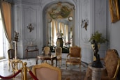 An ornate room with luxurious vintage furniture, intricate molding on the walls, and classical sculptures. The color palette includes soft whites, golds, and browns with elegant chandeliers contributing to the space. Large windows with flowing curtains allow natural light to enter, enhancing the opulence of the room. Decorative wall-mounted lights, a round table with vases, and a plush rug complete the rich interior decor.
