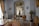 An ornate room with luxurious vintage furniture, intricate molding on the walls, and classical sculptures. The color palette includes soft whites, golds, and browns with elegant chandeliers contributing to the space. Large windows with flowing curtains allow natural light to enter, enhancing the opulence of the room. Decorative wall-mounted lights, a round table with vases, and a plush rug complete the rich interior decor.