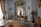 An ornate room with luxurious vintage furniture, intricate molding on the walls, and classical sculptures. The color palette includes soft whites, golds, and browns with elegant chandeliers contributing to the space. Large windows with flowing curtains allow natural light to enter, enhancing the opulence of the room. Decorative wall-mounted lights, a round table with vases, and a plush rug complete the rich interior decor.