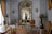 An ornate room with luxurious vintage furniture, intricate molding on the walls, and classical sculptures. The color palette includes soft whites, golds, and browns with elegant chandeliers contributing to the space. Large windows with flowing curtains allow natural light to enter, enhancing the opulence of the room. Decorative wall-mounted lights, a round table with vases, and a plush rug complete the rich interior decor.