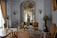An ornate room with luxurious vintage furniture, intricate molding on the walls, and classical sculptures. The color palette includes soft whites, golds, and browns with elegant chandeliers contributing to the space. Large windows with flowing curtains allow natural light to enter, enhancing the opulence of the room. Decorative wall-mounted lights, a round table with vases, and a plush rug complete the rich interior decor.